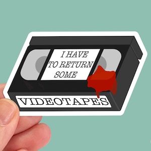 May include: A black and white sticker of a VHS video cassette with red dripping paint. The sticker says "I have to return some videotapes".