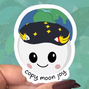 May include: A white sticker with a cartoon moon character wearing a dark blue hat with yellow stars and a green and blue earth on top. The sticker has the text "copy, moon joy."