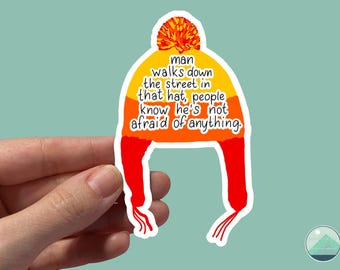 May include: A sticker featuring a colorful winter hat design. The hat is primarily yellow and orange with red accents and a pom-pom. Text on the hat reads: "man walks down the street in that hat, people know he's not afraid of anything."