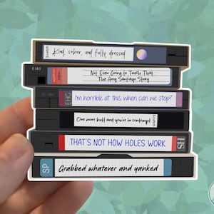 May include: A sticker featuring a stack of VHS tapes with humorous phrases written on them. The tapes are black with white labels and text, including "Kind, sober, and fully dressed" and "That's not how holes work."