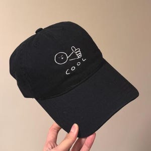 Può includere: Un cappellino da baseball nero con un ricamo bianco che raffigura un semplice omino stilizzato che fa il gesto del pollice in su. Sotto la figura, la parola COOL è ricamata in filo bianco con un carattere informale in stile disegnato a mano.