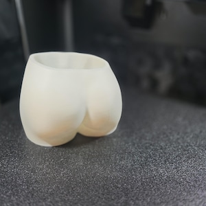 Könnte beinhalten: Ein 3D-gedruckter, cremefarbener Pflanztopf in Form eines Gesäßes. Der Pflanztopf hat eine glatte, abgerundete Form mit einem hohlen Innenraum zur Aufnahme von Pflanzen oder anderen Gegenständen. Der Pflanztopf steht auf einer dunklen, strukturierten Oberfläche.