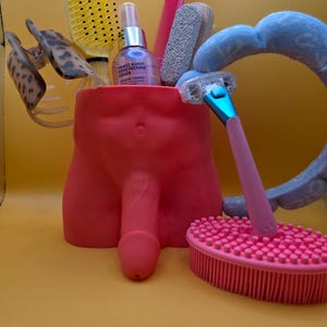 May include: A red, phallic-shaped container holding various grooming tools. Items include a yellow brush, pink comb, razor, and a pink brush. A bottle of toner is also visible. The background is a solid yellow.