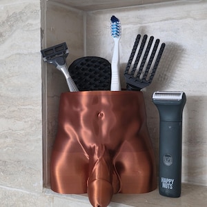 May include: A copper-colored, anatomical-shaped bathroom organizer. It holds a toothbrush, razor, and other grooming tools. A black electric shaver with the text "HAPPY NUTS" is next to the organizer. The organizer is in a tiled shower niche.