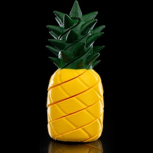 Puede incluir: Una escultura de piña impresa en 3D con un cuerpo amarillo brillante y una parte superior verde y puntiaguda. La piña está segmentada con cortes diagonales, creando una apariencia texturizada. El fondo es negro sólido, resaltando los colores vibrantes.