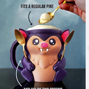 May include: A novelty mug shaped like a cartoon bat, with purple, pink, and tan coloring. The mug has a handle and holds two gold spoons. Text on the image reads "FITS A REGULAR PINT" and "AND HOLDS TWO SPOONS". A scoop of ice cream is being placed in the mug.