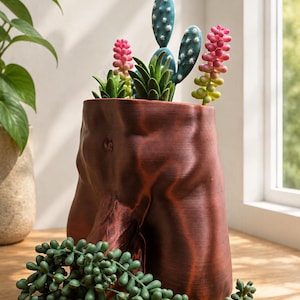 May include: A dark reddish-brown planter shaped like a human torso, containing a variety of artificial succulents including a blue prickly pear cactus, green spiky plants, and tall pink-tipped stalks, with a trailing green string of pearls plant.