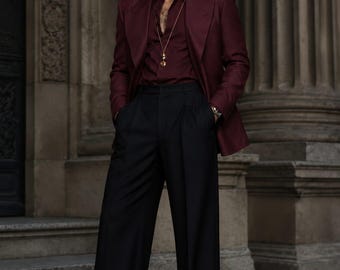 May include: A burgundy blazer worn over a matching burgundy button-down shirt with a gold pendant necklace. The outfit includes high-waisted black wide-leg trousers and dark burgundy leather dress shoes. Dark sunglasses are worn on the face.