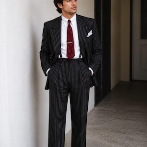 May include: A black pinstripe suit featuring a white dress shirt, a dark red patterned necktie with a gold tie bar, and a white pocket square. The trousers are high-waisted with pleats and cuffs, worn with brown leather dress shoes.