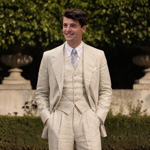May include: A three-piece beige linen suit consisting of a single-breasted jacket with wide lapels, a matching button-front vest, and pleated trousers. The suit is worn with a crisp white collared shirt, a light gray patterned necktie, and a pocket square.