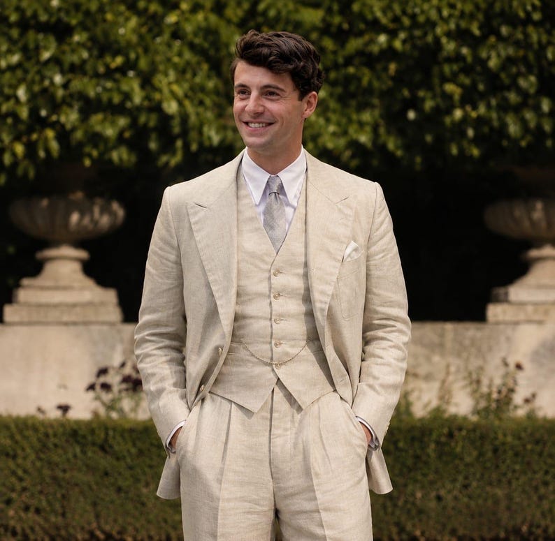May include: A three-piece beige linen suit consisting of a single-breasted jacket with wide lapels, a matching button-front vest, and pleated trousers. The suit is worn with a crisp white collared shirt, a light gray patterned necktie, and a pocket square.