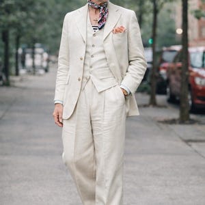 May include: A three-piece cream-colored suit with a matching fedora hat and sunglasses. The suit includes a blazer, vest, and wide-leg trousers. A patterned scarf and pocket square add a pop of color to the ensemble. The outfit is completed with two-tone shoes.
