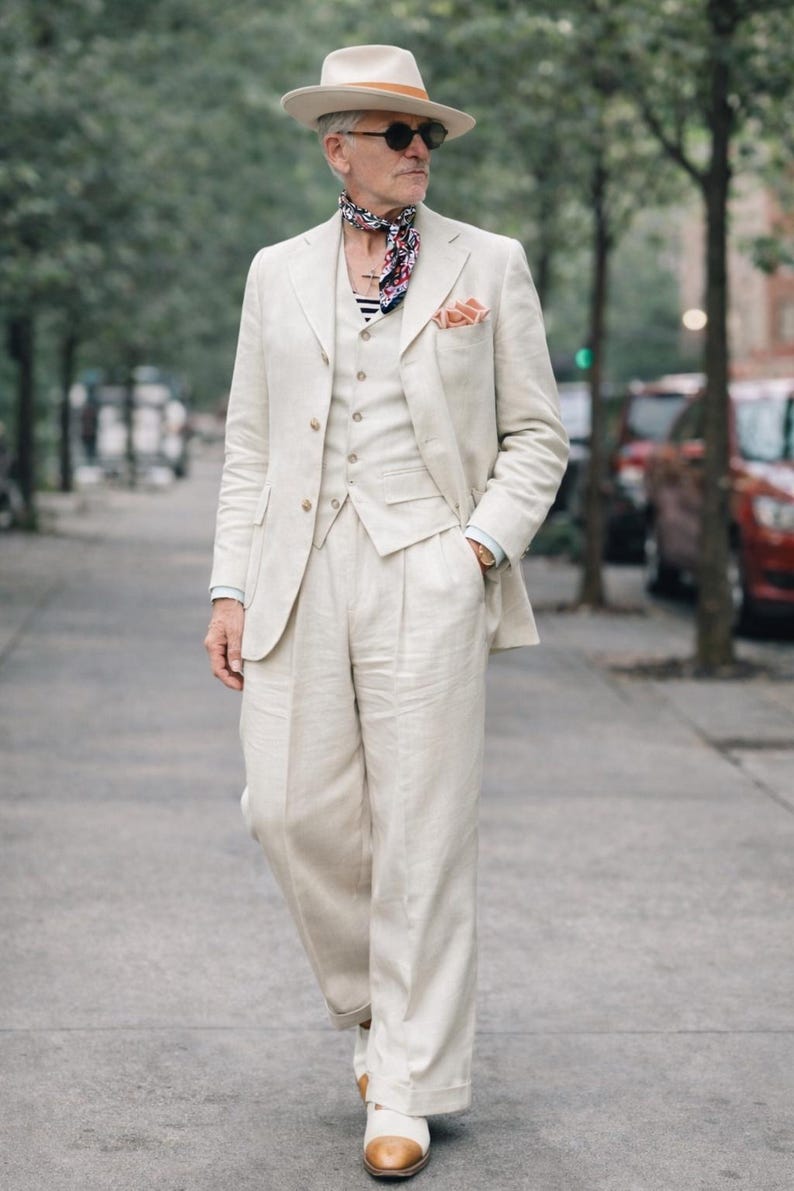 May include: A three-piece cream-colored suit with a matching fedora hat and sunglasses. The suit includes a blazer, vest, and wide-leg trousers. A patterned scarf and pocket square add a pop of color to the ensemble. The outfit is completed with two-tone shoes.