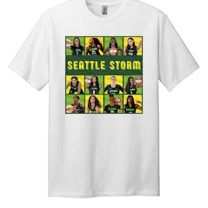 May include: White t-shirt featuring a graphic of the Seattle Storm basketball team. The design includes player images and the team name in green and yellow. The shirt is made of a soft, comfortable material.
