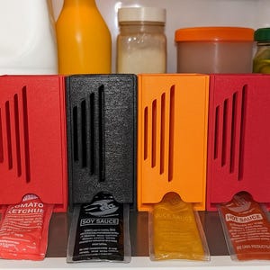 May include: Four colorful condiment holders in a refrigerator. The holders are red, black, orange, and red. Each holder dispenses a packet of sauce: tomato ketchup, soy sauce, duck sauce, and hot sauce. Other items in the fridge include a milk jug and jars.