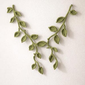 May include: Two green crochet leaf vines are arranged on a white background. The vines are made of yarn and have detailed leaf shapes. The handmade vines are a decorative element.