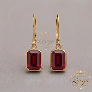 May include: A pair of gold leverback earrings featuring rectangular, emerald-cut deep red gemstones. The earrings have a polished gold setting that secures the stones, which are suspended from a hinged mechanism. The text Craft by Kavya is visible.