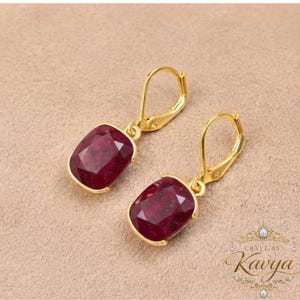 May include: A pair of gold-tone leverback earrings featuring faceted, rectangular deep red gemstones. The earrings have a polished metal finish and a secure hinged closure mechanism, showcasing a classic drop design with vibrant crimson stones.
