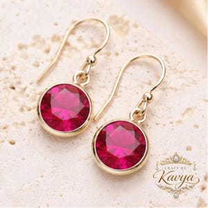 Peut inclure: Une paire de boucles d'oreilles pendantes avec des pierres rondes à facettes d'un rose fuchsia vif, serties dans des montures en métal doré poli. Les boucles d'oreilles sont suspendues à des crochets dorés. Le logo Craft by Kavya est visible.