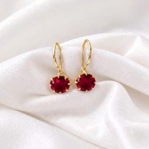 May include: A pair of gold drop earrings featuring round, faceted deep red gemstones. Each earring is suspended from a curved gold wire hook, with the circular red stone held securely in a prong setting, showcasing a polished metallic finish.