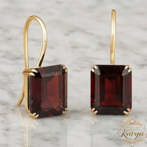 May include: A pair of gold drop earrings featuring large, rectangular, deep red garnet gemstones. Each stone is held by four prongs in a gold setting with a curved wire hook for attachment. The design is symmetrical, showcasing the dark red facets.