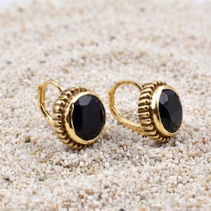 May include: A pair of gold-coloured leverback earrings featuring large, round, black gemstones. The earrings have a textured, ridged border around the black stone. The earrings are displayed on a bed of small, light-coloured pebbles.
