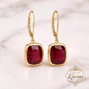 May include: A pair of gold leverback earrings featuring square-cut, deep red gemstones. The earrings have a polished gold finish and a secure hinged closure mechanism. The design is simple, highlighting the vibrant red colour of the stones.