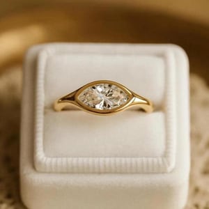 Könnte beinhalten: Ein goldener Ring mit einem Diamanten im Marquiseschliff, der in einer Lünettenfassung gefasst ist. Der Ring wird in einer weißen Samtringbox präsentiert. Der Diamant funkelt und reflektiert das Licht. Das Band des Rings ist aus poliertem Gold.