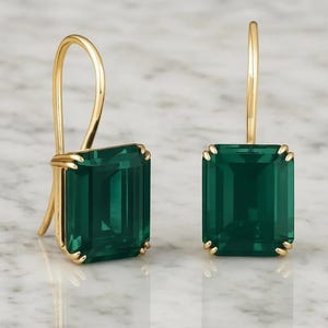 May include: A pair of drop earrings featuring rectangular emerald-cut green stones set in polished yellow gold. Each stone is secured by four prongs and hangs from a simple gold hook, showcasing a deep, vibrant green colour and clear facets.