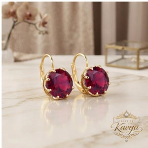 May include: A pair of gold leverback earrings featuring round, faceted deep red stones held by six prongs. The earrings have a polished gold finish and a classic circular design, showcasing a vibrant crimson hue that catches the light.
