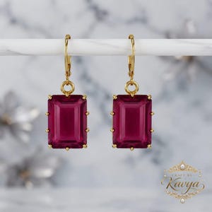 May include: A pair of gold-toned leverback earrings featuring rectangular, deep magenta-coloured faceted stones. The earrings hang from a horizontal bar, and a gold logo with the text Craft by Kavya is visible in the bottom right corner.