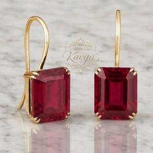 May include: A pair of gold drop earrings featuring large, rectangular, deep red faceted gemstones. Each stone is secured by four prongs in a gold setting with a curved hook wire. The text "Craft by Kavya" is visible in the centre background.