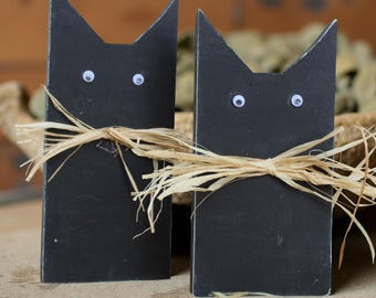 May include: Two black cat-shaped wooden decorations with googly eyes and raffia bows. The cat figures have pointed ears and are standing upright. The background is a blurred mix of brown and tan.