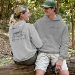 May include: Two people wearing matching gray hoodies with text. The hoodie on the left has text on the back, while the hoodie on the right has text on the front. They are sitting on a log in a wooded area.