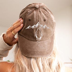 May include: A brown baseball cap with a distressed finish featuring a white embroidered graphic of a mountain range and a penguin standing in the center. The cap has a structured crown, a curved brim, and eyelets for ventilation on the top panels.