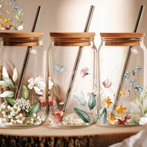 May include: Three clear glass jars with wooden lids and metal straws. Each jar is decorated with watercolor floral designs in white, pink, blue, and yellow. The jars sit on a wooden slice, suggesting a nature-inspired theme.