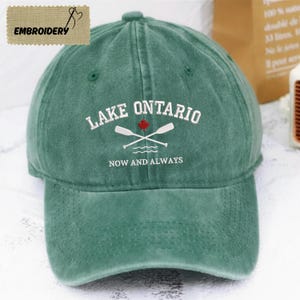 May include: A forest green baseball cap featuring white embroidered text that reads LAKE ONTARIO and NOW AND ALWAYS. The design includes a small red maple leaf icon positioned above two crossed white oars with wavy lines underneath.