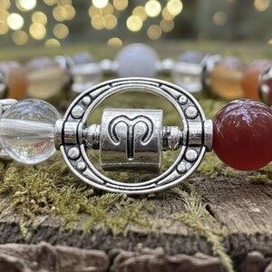Könnte beinhalten: Ein Armband mit Perlen und einem silbernen Widder-Sternzeichen-Anhänger. Das Armband enthält klare, orangefarbene und rote Edelsteinperlen. Der Widder-Anhänger ist zentriert, umgeben von einem silbernen ovalen Rahmen, vor einem moosigen Hintergrund.