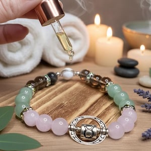May include: A beaded bracelet with pink, green, and brown stones, and a silver Taurus charm. A dropper dispenses oil above the bracelet. The background includes candles, towels, and stones, creating a spa-like atmosphere.