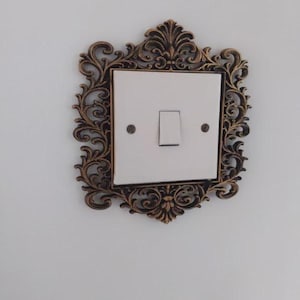 May include: An ornate light switch cover with a bronze-coloured frame. The frame surrounds a white switch plate with a single toggle switch. The design features intricate scrollwork and floral motifs, adding a touch of elegance to the wall.