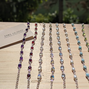 Peut inclure: Plusieurs bracelets en pierres précieuses avec des chaînes en argent sont présentés. Les bracelets présentent diverses pierres colorées, notamment violettes, rouges et bleues, exposées sur une surface en bois. La photo comprend une carte de visite avec le texte "EDEL GLANZZ EDELSTEIN SILBER SCHMUCK".