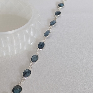 Könnte beinhalten: Ein Silberkettenarmband mit neun ovalen, dunkelblauen, marmorierten Steinen, die in einzelnen Silberfassungen eingefasst sind. Das Armband verfügt über einen silbernen Karabinerverschluss an einem Ende und einen kleinen silbernen Ring am anderen.