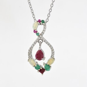Può includere: Una collana con pendente in argento con un design a simbolo dell'infinito. Il pendente è adornato con una varietà di pietre preziose, tra cui un grande rubino a forma di goccia, smeraldi, opali e piccoli diamanti. La collana è appesa a una delicata catena d'argento.