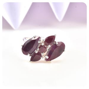 May include: A silver ring featuring a cluster of deep red gemstones. The gemstones are pear and oval-shaped, arranged in a floral-like pattern. The ring is set against a white background, highlighting the vibrant color of the stones.