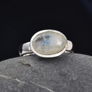 Può includere: Un anello in argento con un cabochon di pietra di luna di forma ovale, incastonato in una montatura in metallo lucido. La gemma presenta un aspetto bianco lattiginoso e traslucido con una sottile iridescenza blu, montata su una fascia in argento.