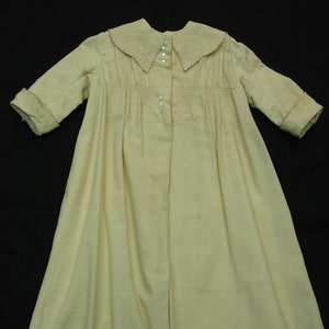 May include: A pale yellow, long-sleeved baby gown. The gown features a Peter Pan collar with embroidered detailing, a smocked bodice, and small pearl buttons. The sleeves have cuffs with embroidered trim. The gown is made of a lightweight fabric.