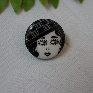 May include: A round, black and white button featuring a stylized face. The face has large eyes, a small nose, and a defined lip shape. The hat has a checkered pattern. The button is likely a decorative accessory.
