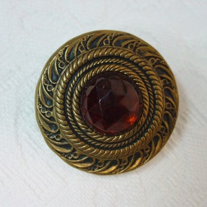May include: A round, gold-toned button with an intricate design and a dark red glass center.
