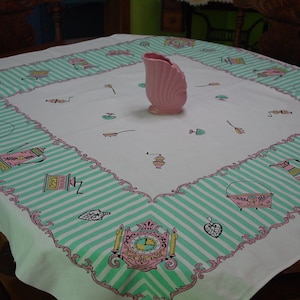 May include: A vintage square tablecloth with a white background and a border of alternating mint green stripes and pink floral designs. The center features small kitchen-themed illustrations and a pink vase.