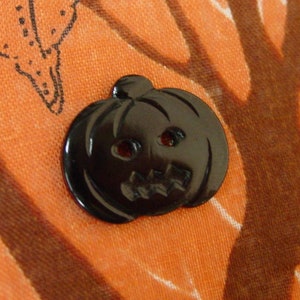 May include: A black plastic pumpkin button with a carved face. The button is on an orange fabric background with a brown tree design.